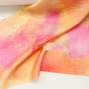 May include: A silk scarf with a watercolour design in shades of pink, orange, and yellow. The fabric is draped, showing the soft texture and colour variations. The scarf's design has a blended, artistic appearance.