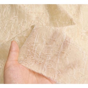 May include: Close-up of a sheer, cream-colored fabric with an intricate floral lace pattern. The fabric is held up, showing its delicate texture and design. The light beige color and detailed embroidery suggest a vintage or elegant aesthetic.