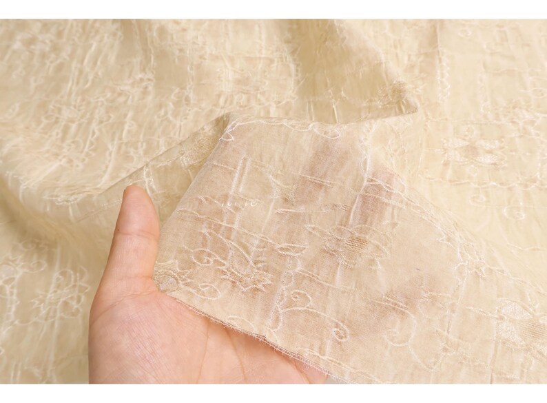 May include: Close-up of a sheer, cream-colored fabric with an intricate floral lace pattern. The fabric is held up, showing its delicate texture and design. The light beige color and detailed embroidery suggest a vintage or elegant aesthetic.