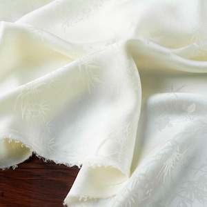 May include: Ivory-colored fabric with a subtle bamboo leaf pattern. The fabric is draped, showing soft folds and a delicate texture. The edges are unfinished, revealing a raw, fringed edge. The overall effect is one of elegance and natural design.