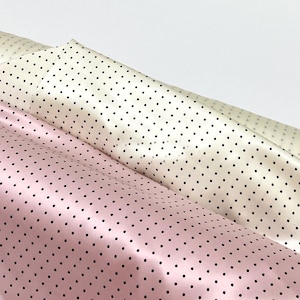 Può includere: Due rotoli di tessuto con un motivo ripetuto di piccoli pois neri. Un rotolo è di un morbido rosa pastello, mentre l'altro è di un bianco crema. Il materiale presenta una texture superficiale liscia e leggermente riflettente.