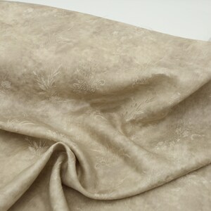 May include: A beige fabric featuring a delicate floral pattern. The material has a soft, textured appearance with light-coloured floral and leaf designs. The fabric is draped, revealing its folds and texture, creating a sense of movement.