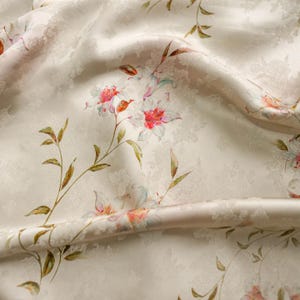 May include: A cream-colored fabric featuring a subtle, tone-on-tone jacquard floral pattern throughout. The material is adorned with a printed design of delicate lilies in shades of soft pink, white, and green, with thin, winding stems.