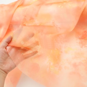 Può includere: Un tessuto trasparente color pesca con motivi ad acquerello nei toni dell'arancione e del giallo. Il tessuto è sollevato, rivelando la sua qualità traslucida. L'effetto complessivo è morbido e arioso, suggerendo un tessuto per abbigliamento o arredamento.