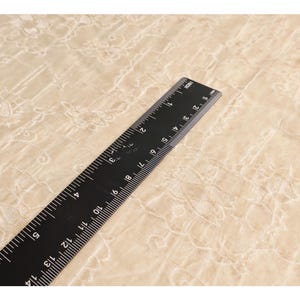 May include: A black ruler with white markings, measuring up to 14 inches, lies diagonally across a beige fabric with a floral pattern. The ruler is made of a matte black material and is clearly marked with inch increments.