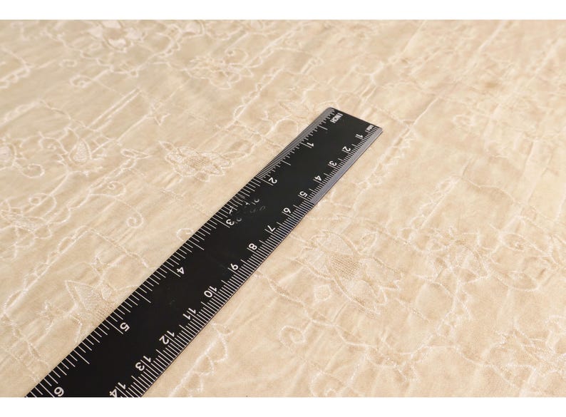 May include: A black ruler with white markings, measuring up to 14 inches, lies diagonally across a beige fabric with a floral pattern. The ruler is made of a matte black material and is clearly marked with inch increments.
