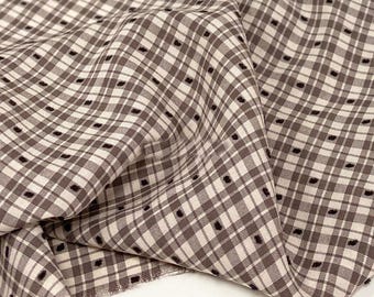 May include: Close-up of a fabric with a brown and cream plaid pattern. The fabric has small, evenly spaced black dots within the plaid squares. The fabric is folded, showing the texture and drape of the material.