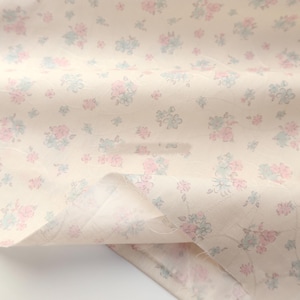 Op de afbeelding: Close-up van een stof met een herhalend bloemenpatroon. De bloemen zijn in tinten roze en blauw, tegen een crèmekleurige achtergrond. De stof is licht gekreukt, wat de textuur en de drapering laat zien. Ideaal voor naaiprojecten.