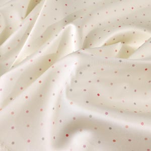 May include: Ivory-colored fabric with a subtle sheen, scattered with small, evenly spaced polka dots in shades of red, pink, and gray. The fabric is draped, creating soft folds and shadows, suggesting a lightweight material like silk or satin.