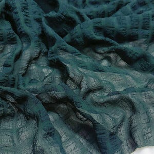 May include: Dark teal fabric with a textured, grid-like pattern. The fabric appears to be lightweight and flowing, with visible folds and shadows. The color is a deep, rich shade of teal, creating a sense of depth and visual interest. The fabric is likely used for apparel or home decor.