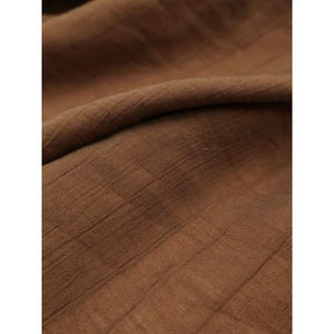 May include: A brown fabric with a visible grid-like texture and a soft, crinkled appearance. The material features a consistent deep earth-tone colour throughout, with subtle folds creating shadows and highlights across the surface of the textile.