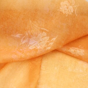 Puede incluir: Primer plano de una tela naranja transparente doblada con un estampado floral. La tela tiene un aspecto suave y texturizado y un brillo sutil. El diseño floral es de un tono naranja más claro, creando un contraste delicado.