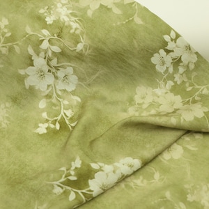 May include: Close-up of green fabric with a floral design. The fabric features a watercolour effect with white flowers and delicate stems. The fabric is folded, showing the texture and depth of the material.