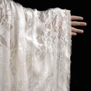 May include: White fabric with a floral pattern. The fabric is held up against a black background. The fabric has a textured appearance and a delicate, sheer quality. The floral design is visible throughout the fabric.