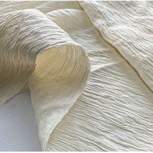 May include: Close-up of a cream-colored fabric with a textured, crinkled surface. The fabric is draped and folded, revealing the interior texture. The material appears soft and lightweight, suitable for clothing or home decor.