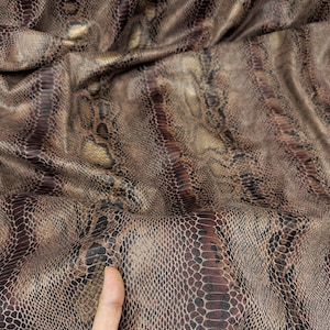 May include: Close-up of a brown and tan snakeskin-patterned fabric. The fabric has a textured surface with a repeating scale design. The color palette includes shades of brown, tan, and a hint of gold.