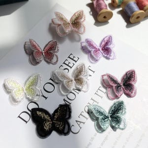 May include: A collection of eight embroidered butterfly patches in various colors including pink, purple, white, green, and black. Each butterfly features intricate thread detailing and a delicate, semi-transparent wing structure. The patches are arranged on a white surface with printed text that reads DO YOU SEE WHAT I SEE.