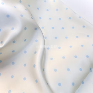 Puede incluir: Tela blanca con un delicado estampado de lunares azul claro. La tela es suave y fluida, con pliegues suaves y un brillo sutil. Los lunares están espaciados uniformemente.