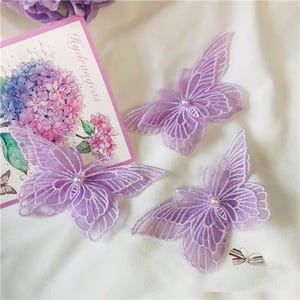 May include: Three purple lace butterfly appliques with pearl accents. The butterflies are delicate and intricate, perfect for adding a touch of whimsy to any craft project.