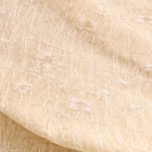 May include: Close-up of a beige fabric with intricate white floral embroidery. The fabric has a delicate, sheer quality, with a subtle sheen. The design features repeating floral patterns within a grid-like structure, creating a textured appearance.