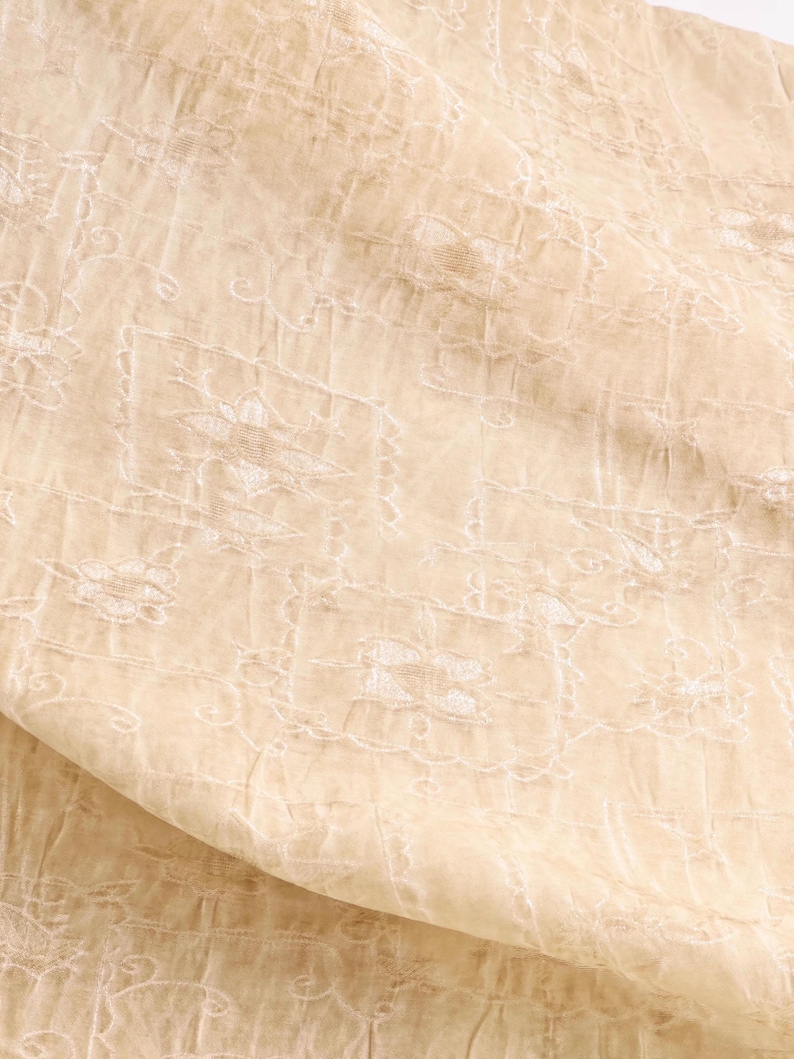 May include: Close-up of a beige fabric with intricate white floral embroidery. The fabric has a delicate, sheer quality, with a subtle sheen. The design features repeating floral patterns within a grid-like structure, creating a textured appearance.