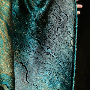May include: Close-up of a teal fabric with a marbled pattern and gold accents. The fabric has a subtle sheen and a textured surface.