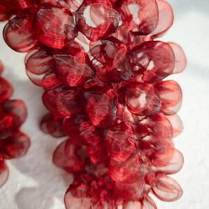 May include: Close-up of a cluster of red, semi-transparent, fabric grape-like shapes. The fabric has a slightly textured appearance, with darker red accents. The shapes are arranged in a cascading, organic form against a white background.