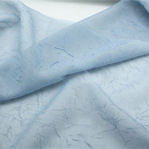 May include: Pale blue fabric with a textured, crinkled appearance. The fabric is folded and draped, showing variations in tone. The material looks lightweight and translucent, possibly for clothing or decorative use.