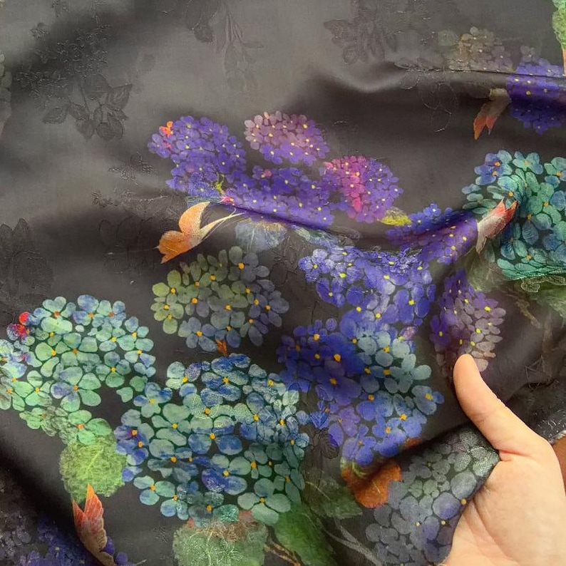 May include: Dark fabric with a floral design. Clusters of blue, purple, and green flowers are set against a dark grey background with embossed floral details. The fabric is held, showing its drape and texture.