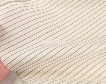 May include: A piece of off-white fabric featuring a pattern of thin, evenly spaced dark brown diagonal stripes. The textile has a visible weave texture and a raw, unfinished edge along the top, displaying a consistent linear design throughout.