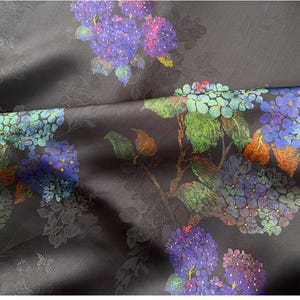 May include: Dark grey fabric with a floral design of purple, blue, and green hydrangea flowers. The material has a slight sheen and an embossed pattern of leaves and flowers. The fabric creates a rich, textured appearance.