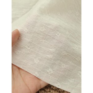 May include: A sheer white fabric featuring a delicate grid pattern formed by thin vertical and horizontal lines. Small, embroidered floral motifs are evenly spaced within the squares of the grid, creating a subtle, textured, and lightweight appearance.