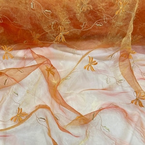 Puede incluir: Primer plano de tela naranja transparente con bordados de flores y hojas. La tela tiene un aspecto fluido y en capas, con diferentes tonos de naranja y toques de blanco. El bordado es de color amarillo dorado.