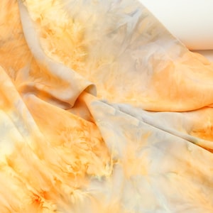 Puede incluir: Primer plano de una tela con un patrón tie-dye en tonos naranja, amarillo y azul claro. La tela está doblada, creando sombras suaves y resaltando la textura y las variaciones de color.