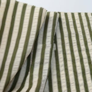 May include: Close-up of a fabric with vertical stripes in olive green and off-white. The fabric is folded, showing the texture and pattern. The stripes are evenly spaced, creating a classic design.
