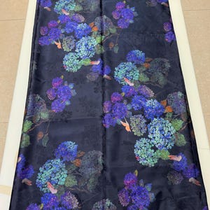 May include: Dark navy fabric with a floral pattern of blue, purple, and green hydrangeas. The design includes small birds, suitable for apparel or home decor.