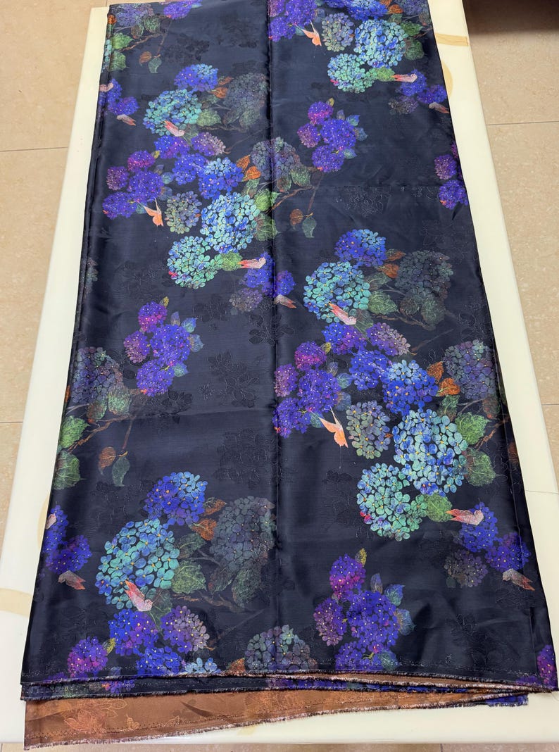 May include: Dark navy fabric with a floral pattern of blue, purple, and green hydrangeas. The design includes small birds, suitable for apparel or home decor.