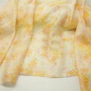 Puede incluir: Una bufanda de tela ligera y semitransparente con un delicado patrón floral disperso en tonos amarillo dorado y rosa pastel suave. El tejido presenta una textura visible y cae en pliegues sueltos y fluidos sobre la superficie.