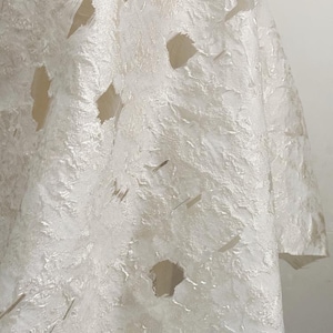 May include: Close-up of a textured, off-white fabric with irregular cut-out shapes. The fabric has a layered, almost paper-like appearance, with a slightly reflective sheen. The overall effect is delicate and artistic, suggesting a garment or decorative textile.