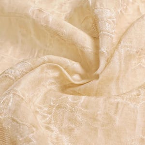 May include: Close-up of a cream-colored fabric with a subtle floral pattern. The fabric is draped, creating soft folds and shadows. The texture appears smooth with a slight sheen, suggesting a delicate material like silk or satin.