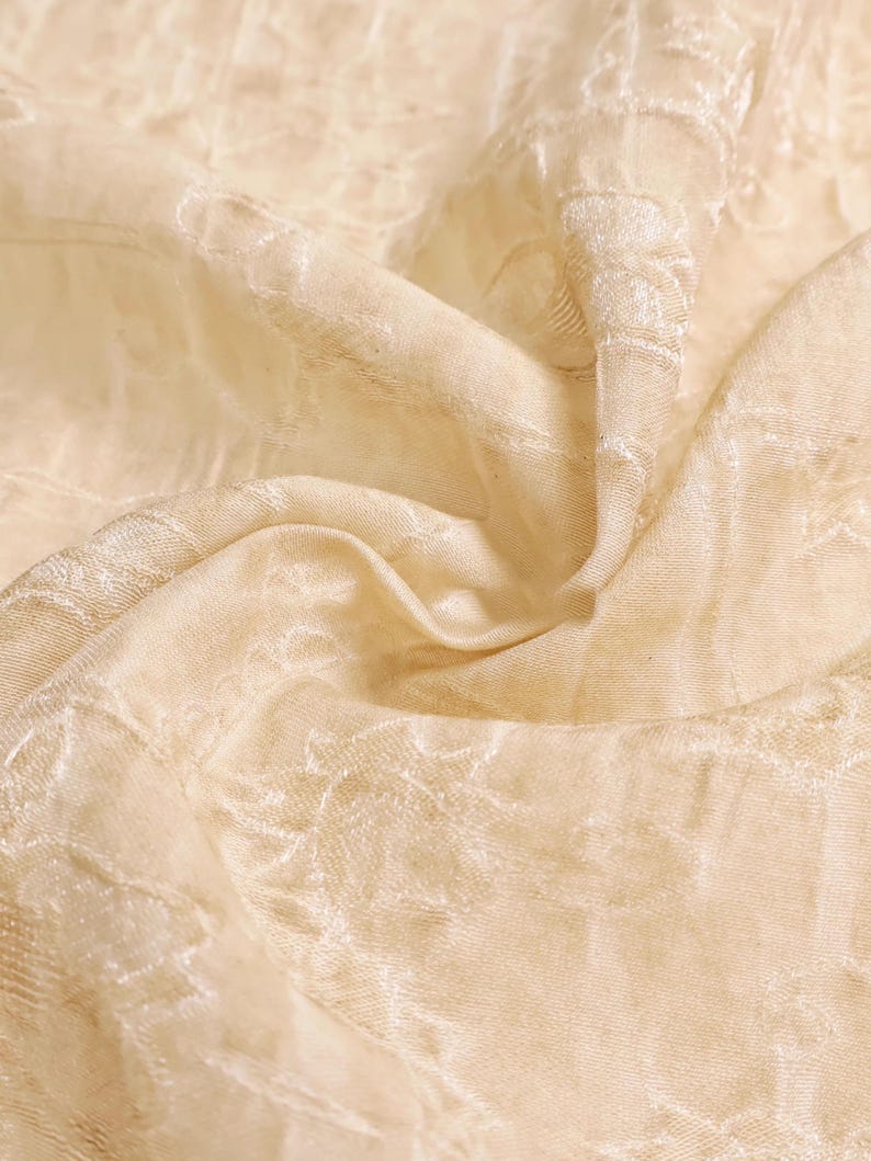 May include: Close-up of a cream-colored fabric with a subtle floral pattern. The fabric is draped, creating soft folds and shadows. The texture appears smooth with a slight sheen, suggesting a delicate material like silk or satin.