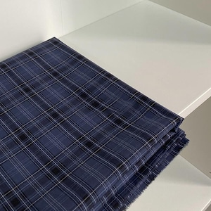 May include: A folded length of navy blue plaid fabric, featuring a grid pattern in black and white. The fabric is neatly stacked on a white shelf, highlighting the pattern. The edges of the fabric are slightly frayed.