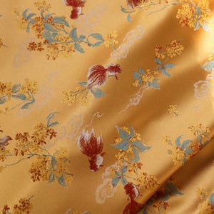 Könnte beinhalten: Nahaufnahme eines goldgelben Stoffes mit einem sich wiederholenden Muster aus roten Fischen, gelben Blumen und blaugrünen Blättern. Der Stoff hat einen leichten Glanz und eine weiche Textur mit Falten und Schatten.