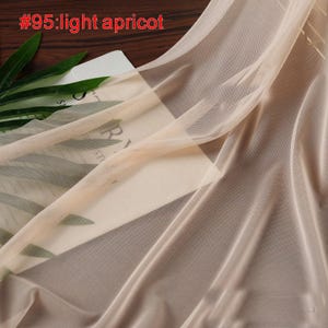 Op de afbeelding: Licht abrikooskleurige transparante stof met een fijne mesh textuur. De stof is gedrapeerd en onthult een glimp van een boek en groene bladeren op de achtergrond. De tekst "#95:light apricot" is zichtbaar linksboven.