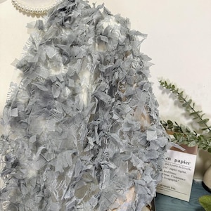 May include: A light gray fabric with a textured, layered design. The fabric is draped over a mannequin, with a pearl necklace visible at the neckline. The fabric appears to be a decorative textile, possibly for apparel or crafts.