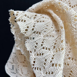 May include: Close-up of a cream-colored, open-weave knitted shawl. The shawl features a delicate, lacy pattern with scalloped edges. The texture is soft and the color is a warm, off-white. The knitted design is visible throughout the fabric.