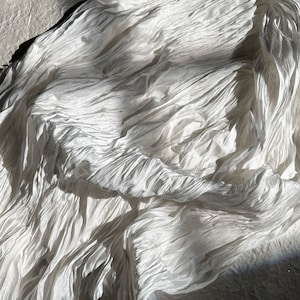 Op de afbeelding: Witte stof met een gekreukelde textuur, wat een zachte en vloeiende uitstraling creëert. De stof is gedrapeerd en gevouwen, waardoor de delicate en luchtige aard zichtbaar wordt.