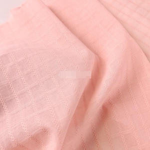 May include: A light pink fabric featuring a subtle, repeating grid pattern created by woven, textured lines. The material has a soft, semi-sheer appearance with folds that highlight the consistent geometric structure of the weave throughout the textile.