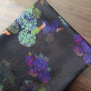 May include: Dark fabric with a floral design. The fabric displays clusters of blue, purple, and green hydrangea flowers. The fabric has a subtle sheen and a dark background, creating a contrast with the colourful flowers.