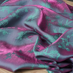 May include: A piece of iridescent fabric featuring a deep magenta and dark grey colour-shifting base. The material is adorned with a repeating pattern of teal green floral blossoms and bamboo stalks, creating a textured and reflective appearance.